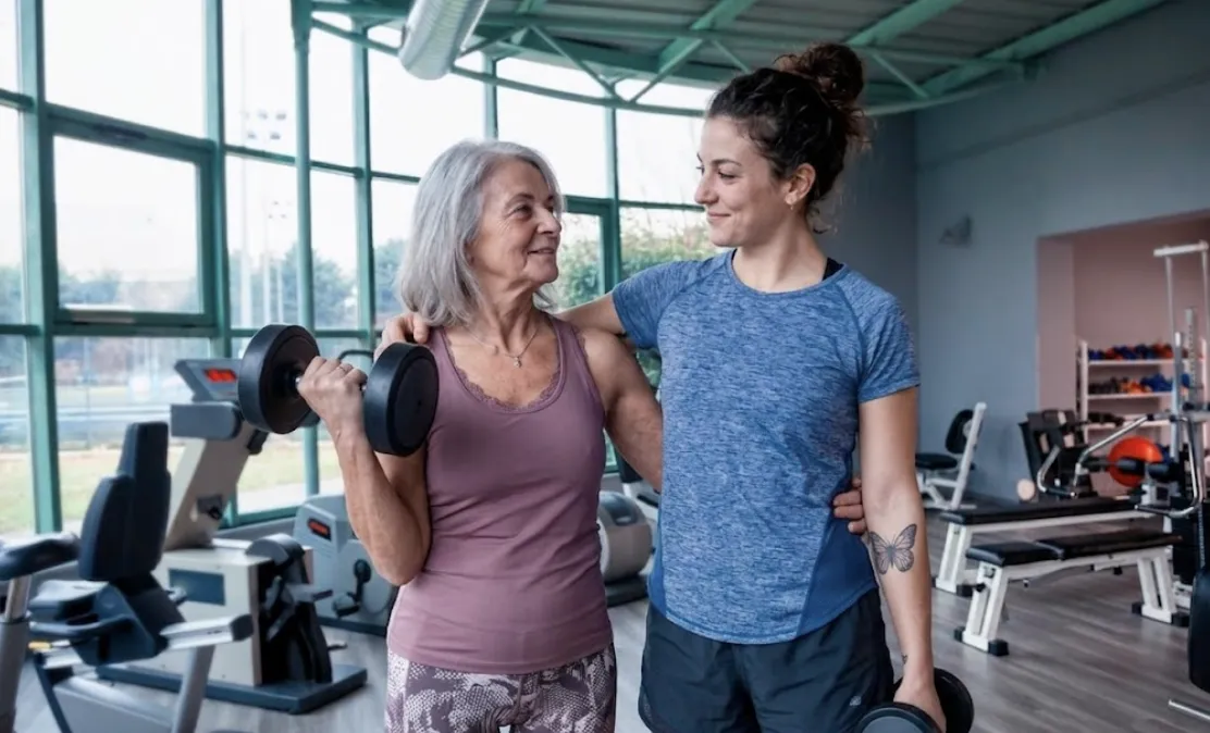 An experienced, young woman personal trainer in Miami's Brickell area, providing specialized, hands-on dumbbell lifting guidance to an active older woman, illustrating how personalized strength training can support fitness, bone density, and muscle mass during the menopause transition. The quiet, modern studio setting emphasizes privacy and expert support for active women over 50.